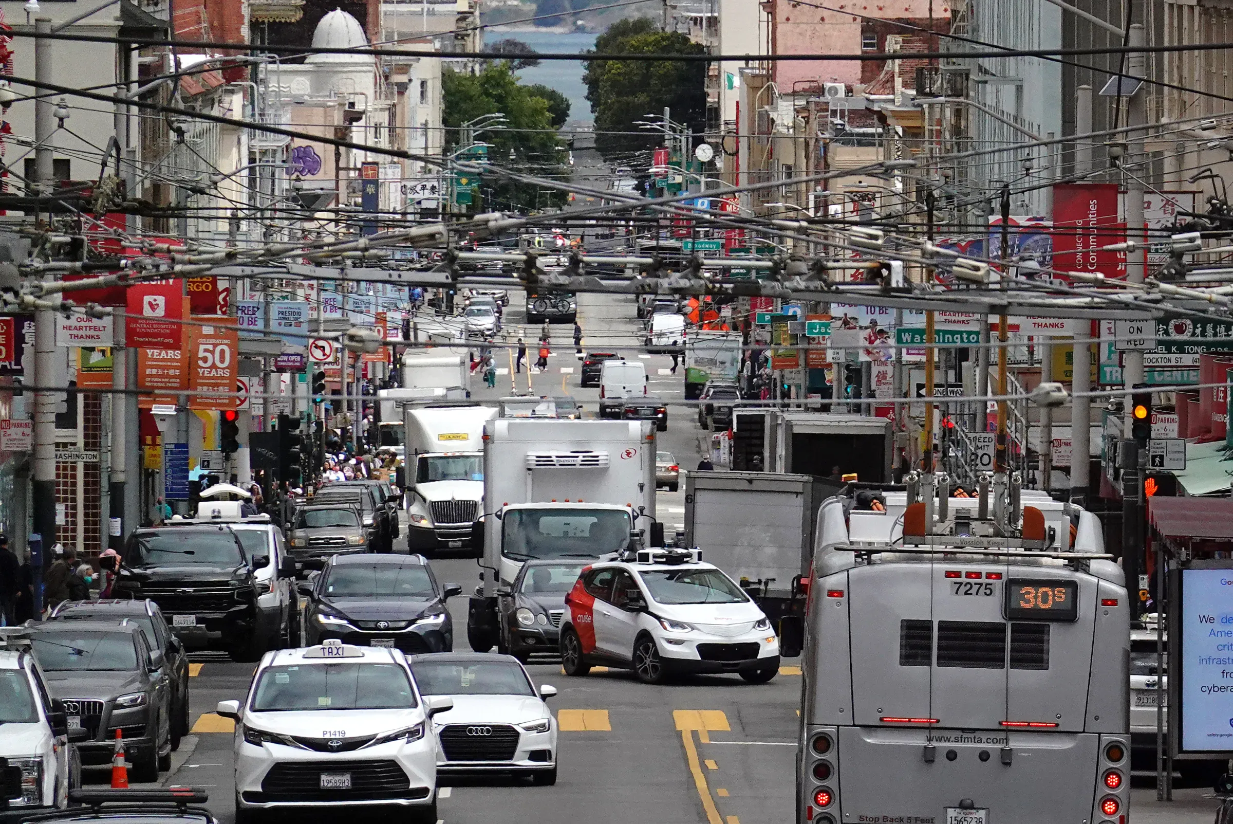 San Francisco city street with Muni bus, autonomous vehicle, and dense urban traffic under overhead trolley wires